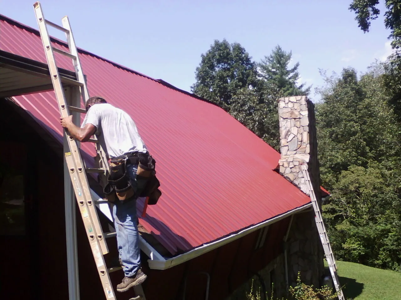 Roofer on a ladder beside a red metal roof and stone chimney — Soffit & Fascia Repair by Roofing Brothers in Pocatello